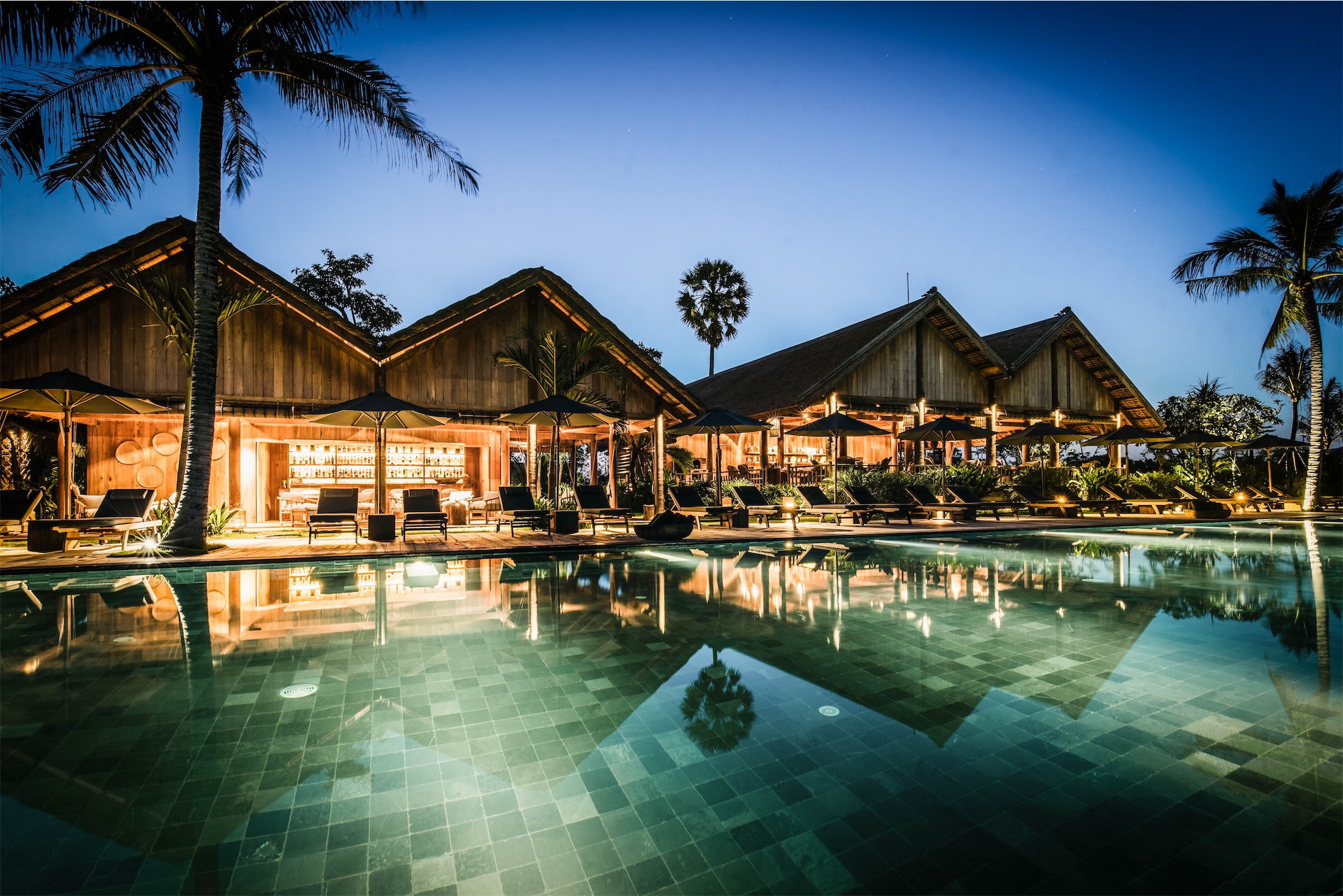 Illuminated pool and wooden pavilions reflected in water at night at Zannier Phum Baitang.