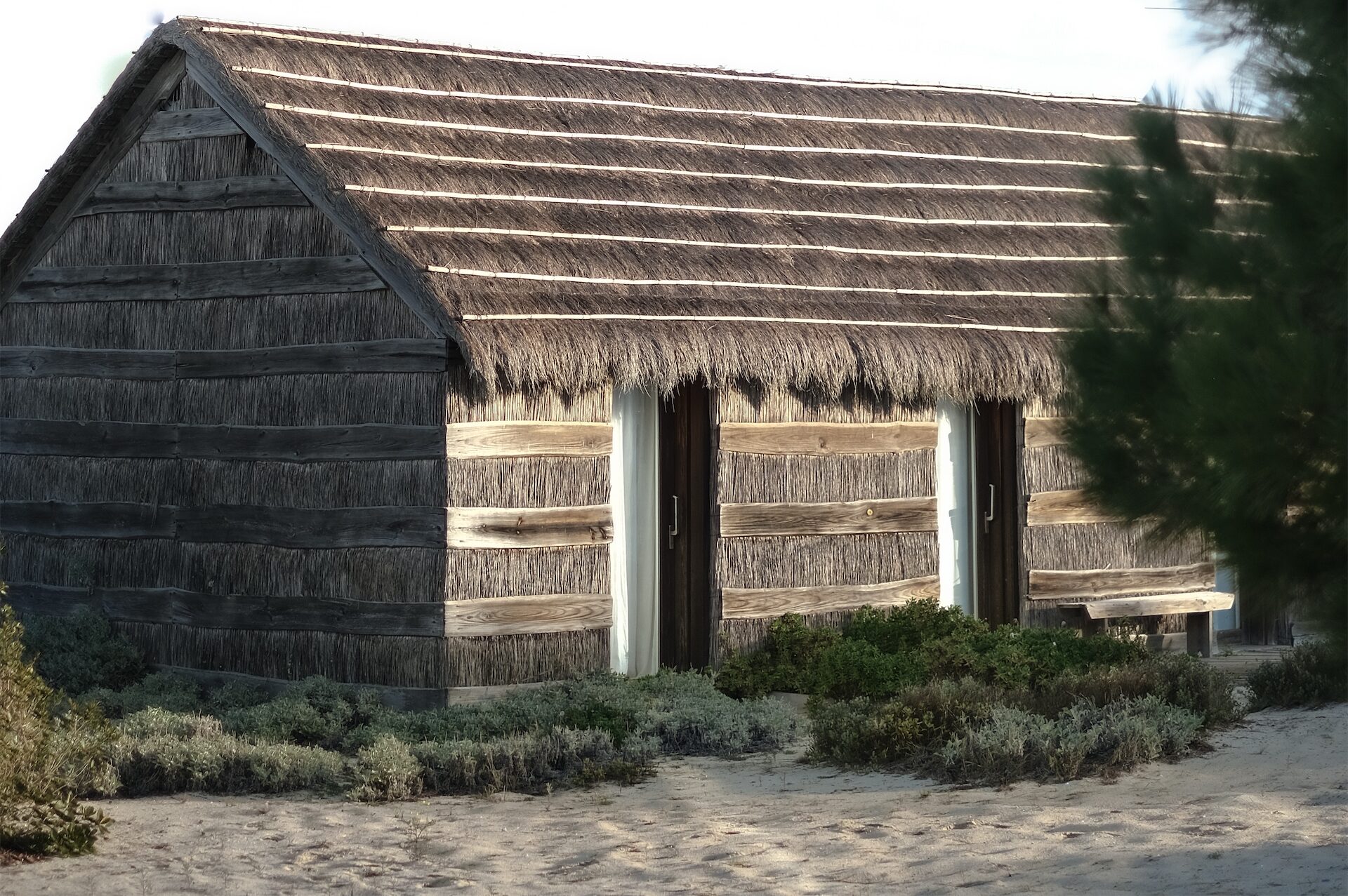 Traditional thatched cottage at Casa na Areia in Comporta, Portugal, surrounded by sand and coastal vegetation.
