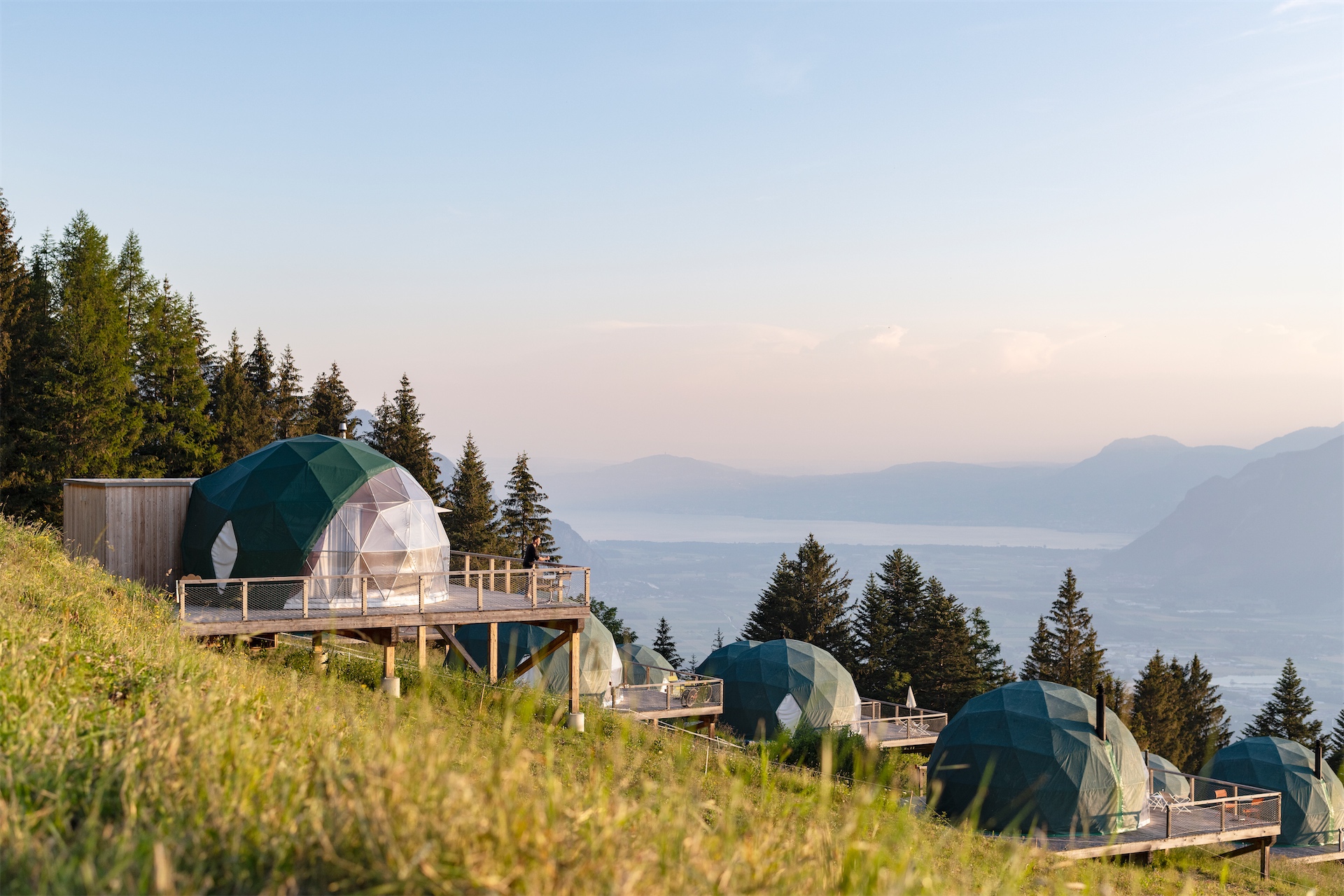 Geodesic pods at Whitepod Original set along a grassy alpine hillside overlooking a lake and valley.