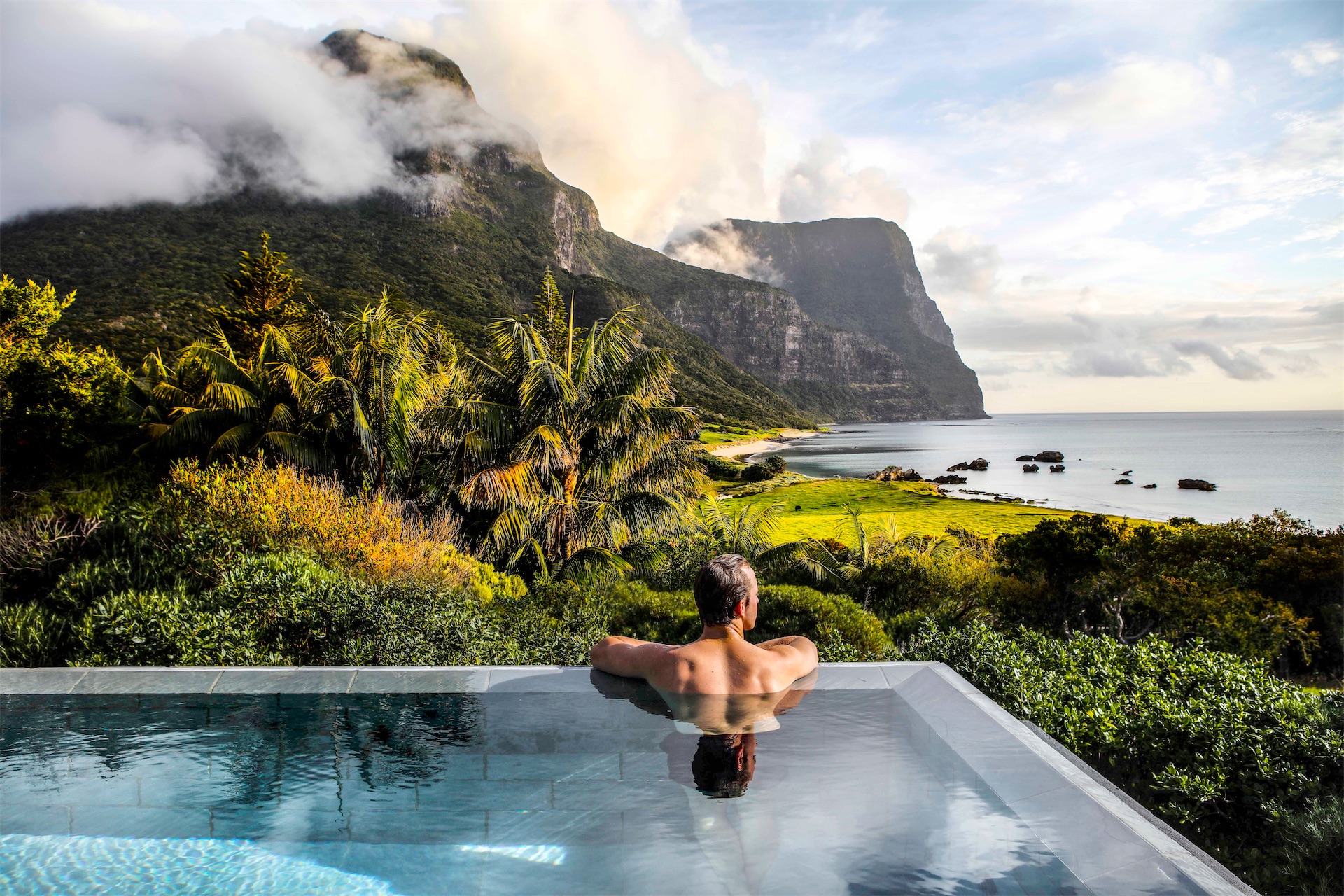 Man relaxing in Capella Lodge infinity pool overlooking Lord Howe Island beach, lagoon, and cloud-capped volcanic peaks at dawn