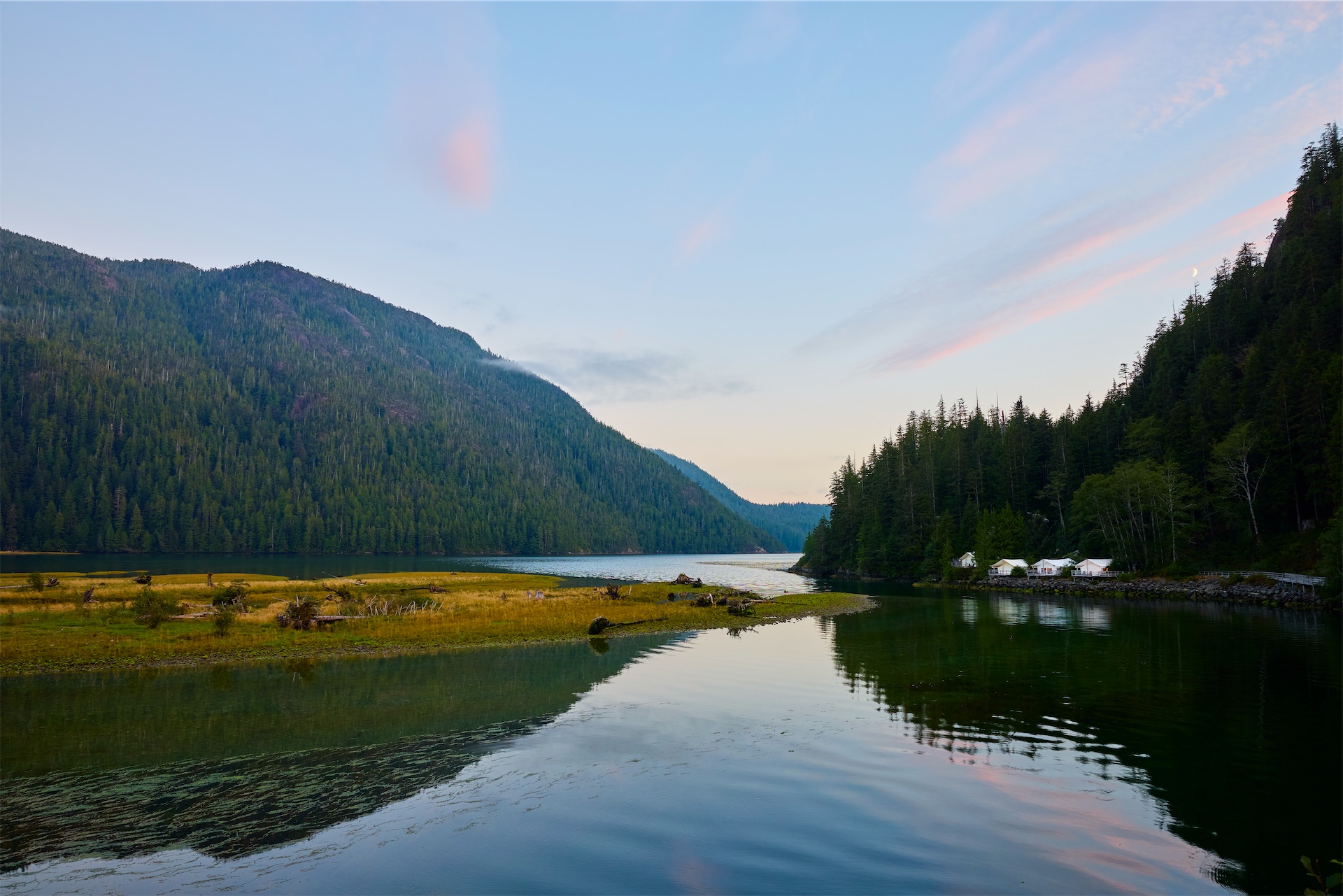 Calm inlet at dusk near Clayoquot Wilderness Lodge with forested mountains, golden grasses, and white lodge structures reflected in still water