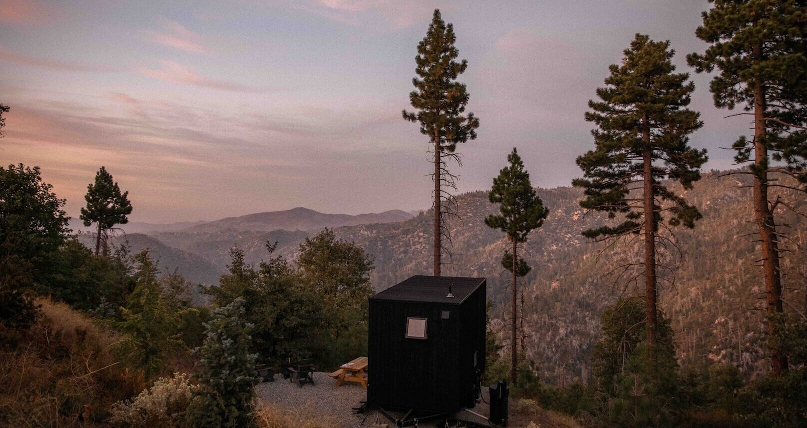 Minimalist black cabin overlooking forested mountains at sunset, surrounded by tall pine trees