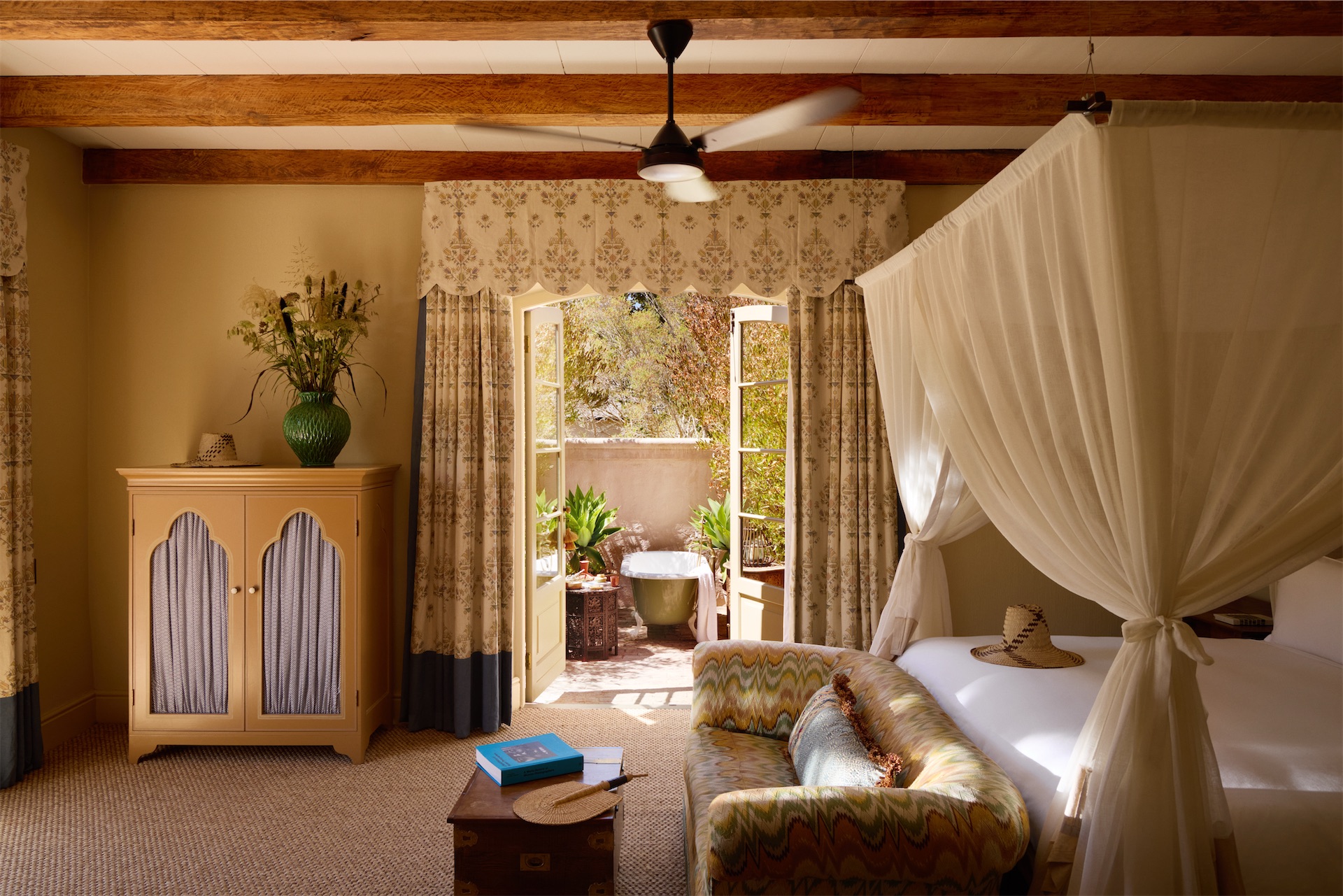 Sterrekopje Farm bedroom with canopy bed, patterned curtains, and French doors opening to a private outdoor bathtub amid lush greenery.
