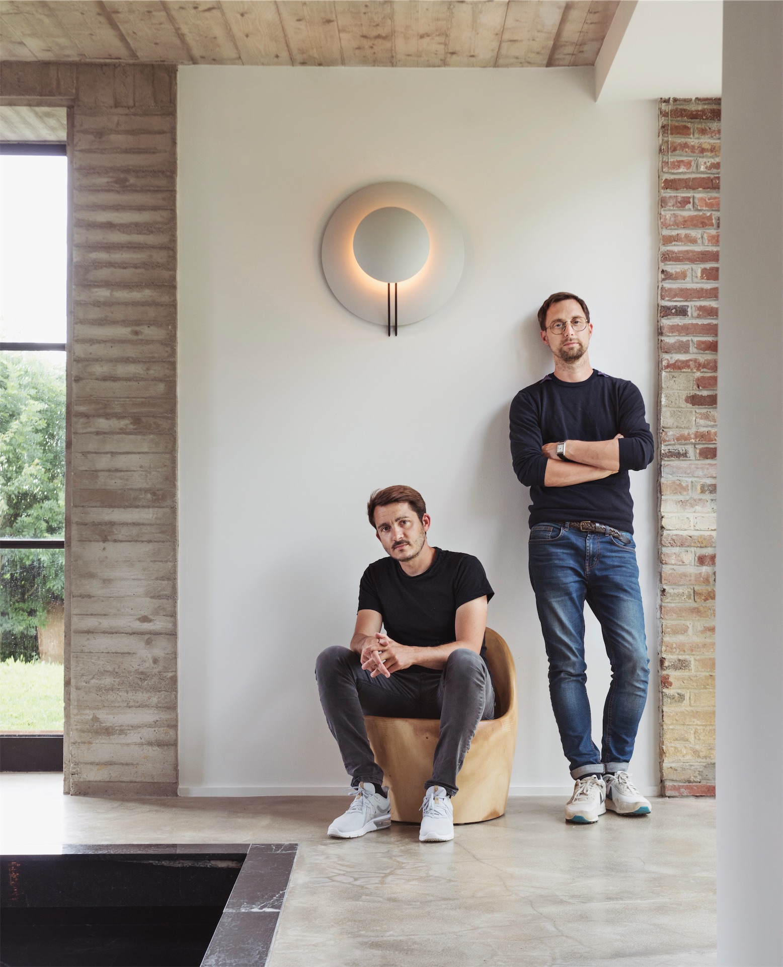 Two men, founders of Maison Ceronne, posed inside the Le Perche country house — one seated in a sculptural chair, one standing against a white wall.