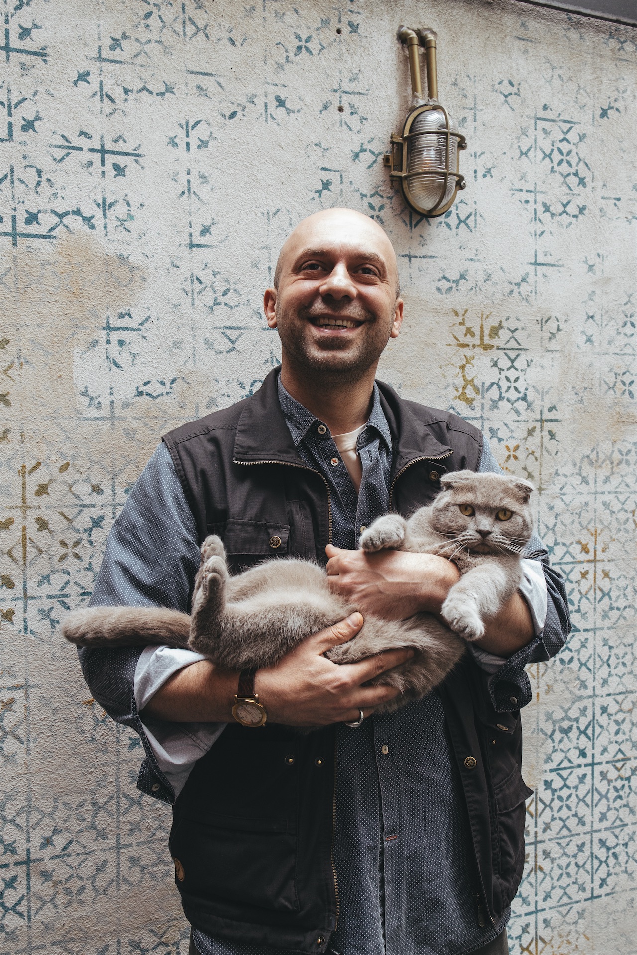 Smiling man Yusuf Çakır holds a grey Scottish Fold cat against a stencil-patterned wall at The Yard Hotel Istanbul.